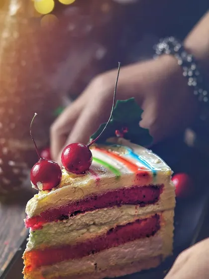A slice of colorful rainbow cake being served, with a human hand adding a personal touch. The warm, festive lighting and soft focus on the Christmas decor in the background tell a story of holiday indulgence.