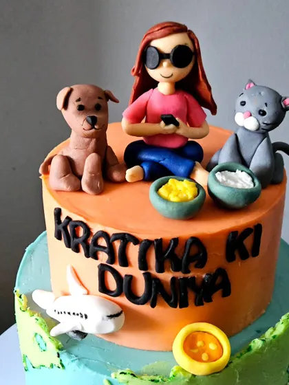 A close-up of the top tier of the "Krattika Ki Duniya" cake. This view highlights the handmade figurines of Krattika with her dog and cat, showcasing the personal details that make these toppers special.