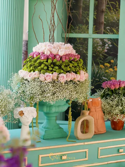 A large floral arrangement featuring tiers of pink roses and baby's breath, set in a mint-green urn. This piece is a beautiful example of structured yet romantic floral design.