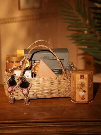 A rustic and elegant Diwali hamper in a wicker basket. This gift includes jars of gourmet food, diyas, and a pair of beautiful mirror-work earrings.