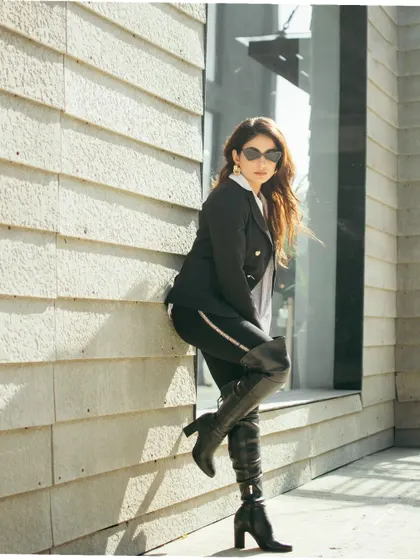 A full-length shot showcasing a powerful pose against a modern, textured wall. The strong lines of the building and the thigh-high boots create a bold, high-fashion statement.
