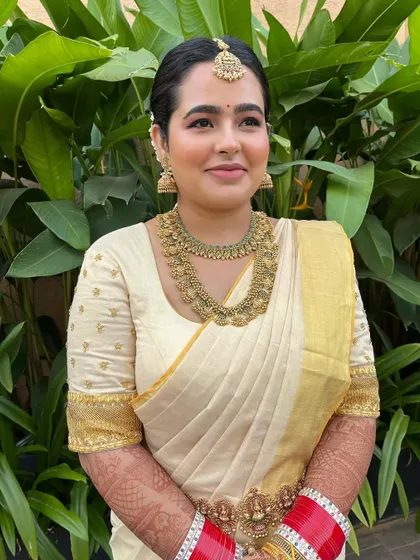 A full-frontal portrait of the bride in her golden-white Mundu saree. The makeup is minimal and fresh, perfect for an early morning ceremony.