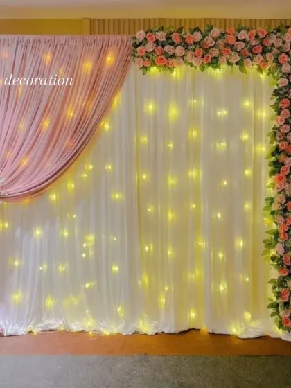 For a soft and elegant look, I use fabric drapes with fairy lights behind them. This backdrop features white and pink drapes, an artificial flower border, and a matching floral arrangement on a stand, ideal for engagements or baby showers.
