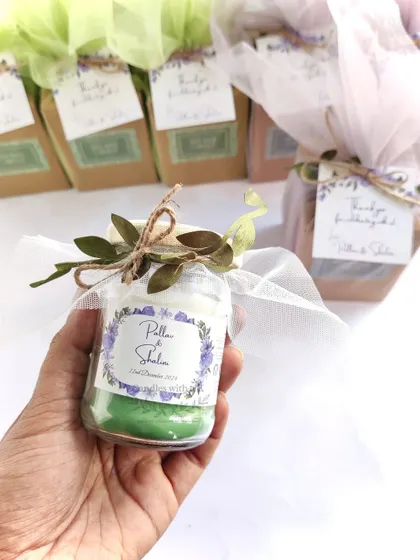 A hand holding a fully packaged wedding favor, a green layered candle with a custom "Pillai & Shalini" sticker, white net wrap, and leafy tie.