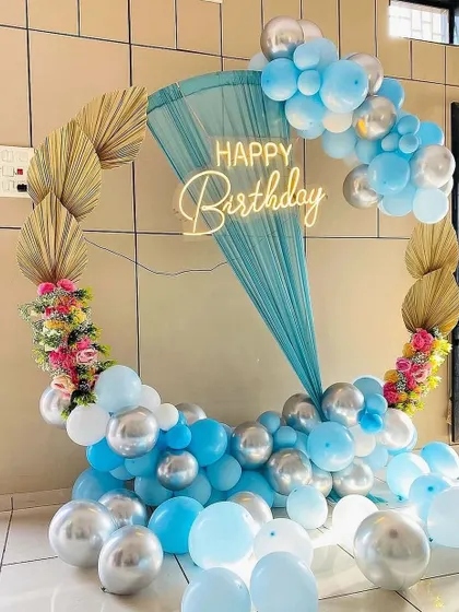 A unique birthday backdrop featuring a circular frame with blue and silver balloons, dried palm leaves, and a sheer blue drape.