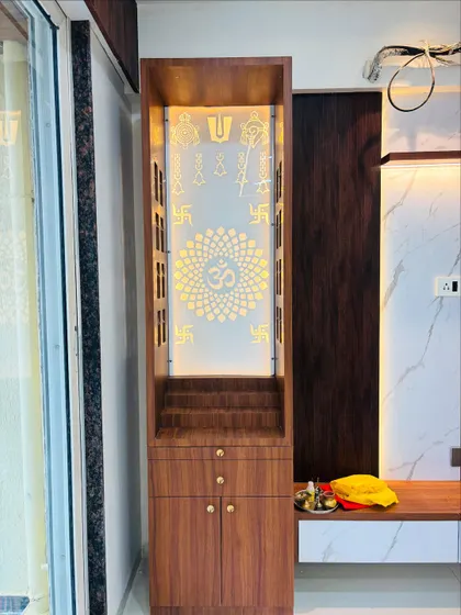 A close-up of a mandir integrated next to a TV unit. The backlit panel features traditional Hindu symbols, and the wooden finish matches the living room furniture for a cohesive look.