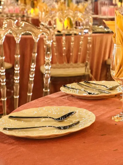 A beautiful table setting featuring textured plates, modern black flatware, and elegant transparent Napoleon chairs. The peach-colored tablecloth adds a soft, warm touch.