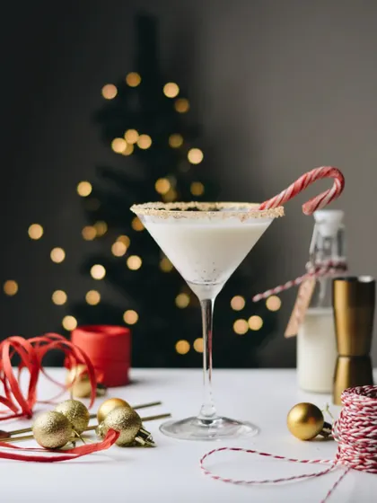 A classy, festive eggnog-style cocktail with a candy cane garnish. The clean, bright styling against the bokeh background gives it a sophisticated holiday party vibe.
