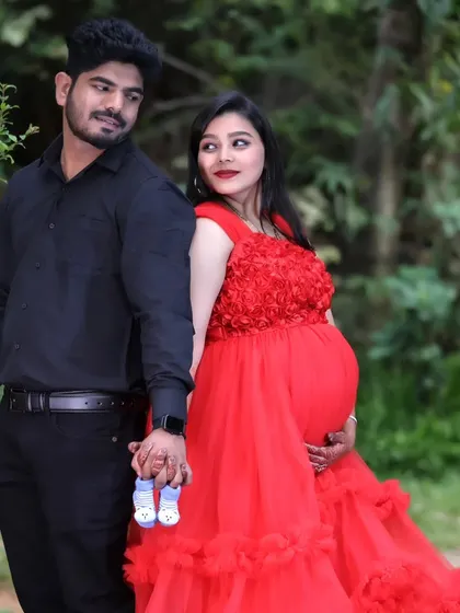 A romantic couple's maternity shoot in a garden. The woman wears a red ruffled gown, and they hold a pair of tiny baby shoes.