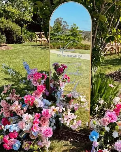 A modern and glamorous welcome sign made of a gold-framed mirror, surrounded by colorful flowers.