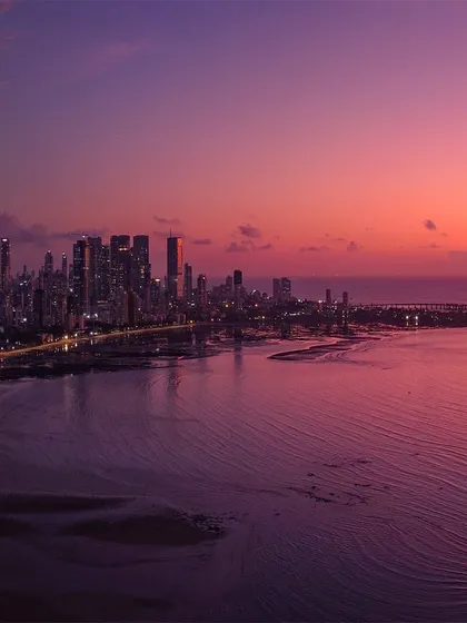 A panoramic view of the Mumbai coastline and the Bandra-Worli Sea Link, bathed in the purple and pink light of a dramatic sunset.