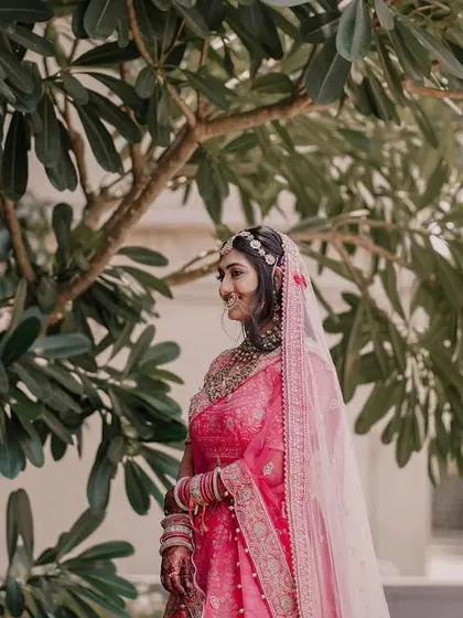 A serene bridal portrait in a garden setting. The bride, dressed in a beautiful pink lehenga, stands under a tree, creating a natural and peaceful image.
