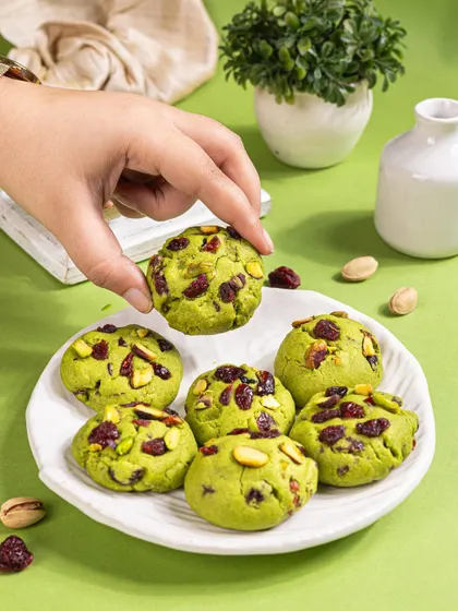 A lifestyle shot of someone picking up a pistachio cranberry cookie from a plate, adding a human touch.