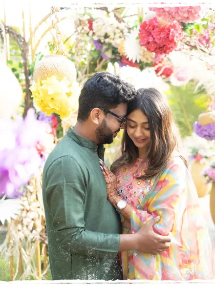 A couple embraces amidst a colorful, floral decor setup for their Mehendi. Their closed eyes and gentle smiles show a moment of quiet connection and happiness.