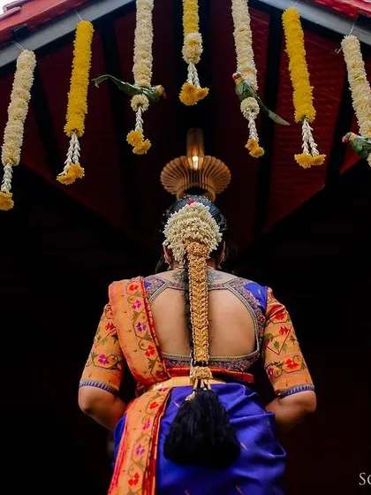 A shot from behind the bride, focusing on her elegant blue saree, intricate blouse, and traditional hairstyle against a backdrop of hanging flowers.