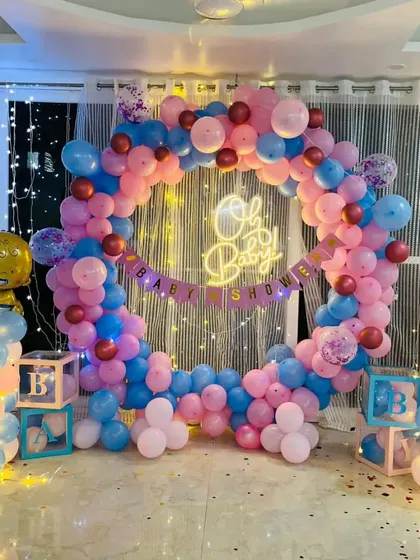 A full room view of a beautiful baby shower decoration. The centerpiece is a large balloon ring in pink and blue, illuminated by fairy lights and a central "Oh Baby" neon sign.