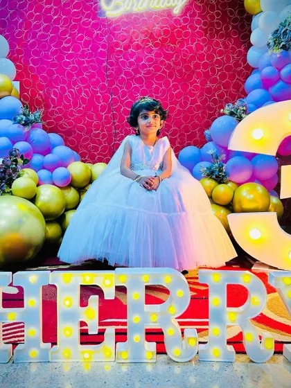 A full-length shot of the birthday girl in her beautiful white dress, standing in front of her name in marquee letters. The vibrant decor makes the photo pop.