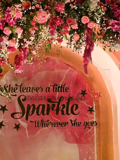 A close-up of the "She leaves a little sparkle" sign at an Aqeeqah ceremony. The sign is beautifully framed by a lush arch of pink and purple flowers.