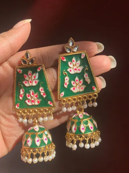This pair of dark green Meenakari jhumkas features a hand-painted lotus motif and pearl accents. The trapezoid shape of the stud adds a modern touch to the traditional design.