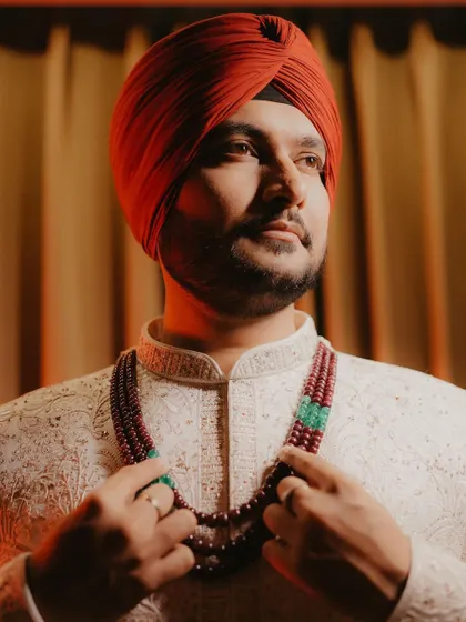 A handsome portrait of a Sikh groom in his traditional white sherwani and red turban. The focus is on his regal attire, including the beaded necklace.
