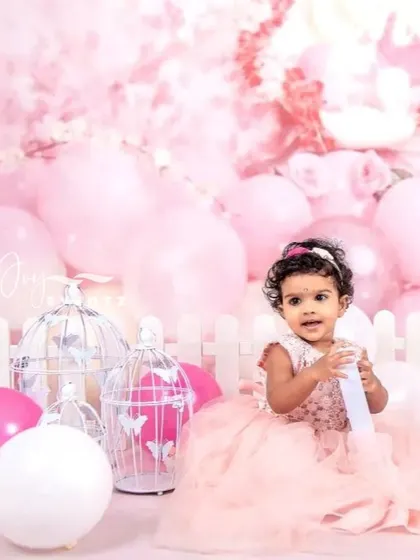 A dreamy and elegant cake smash photography session for a baby girl. Dressed in a beautiful pink tulle gown, she is surrounded by a cloud of pink balloons and decorative birdcages, making for a truly special portrait.