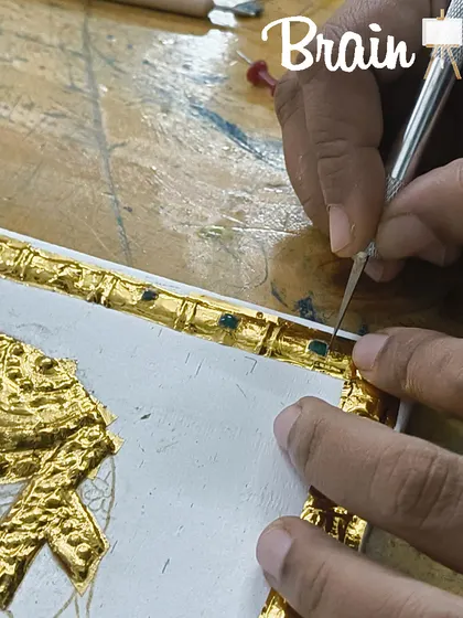 The intricate process of setting stones into the gold foil border of a Tanjore painting. This detailed work adds a three-dimensional, jeweled effect to the final piece.