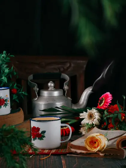 A beautiful still life composition with a vintage tea set, an open book, and flowers against a dark background. The single wisp of steam adds a touch of life to this moody, classic scene.