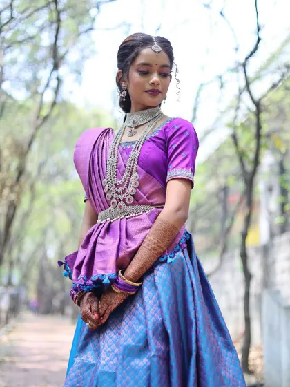 A full-length outdoor portrait of the bride in a beautiful blue and purple traditional half saree. The natural light and green background make the colors of her outfit pop.