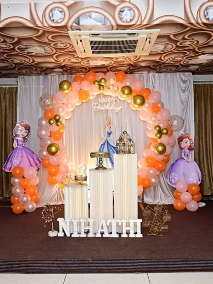 A 'Sofia the First' themed birthday party decoration. This setup includes character standees, an orange and gold balloon arch, and white plinths for the cake and desserts.