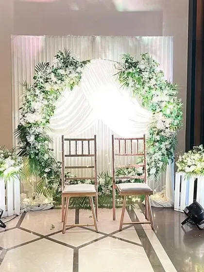 A charming and rustic Nikaah or engagement seating area. The setup features two wooden chairs under a draped arch adorned with white flowers and greenery.