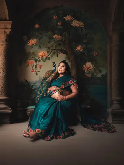 A beautiful portrait of a mom-to-be in a teal saree, seated against a hand-painted backdrop. The peacock adds a touch of traditional artistry to this unique studio session.