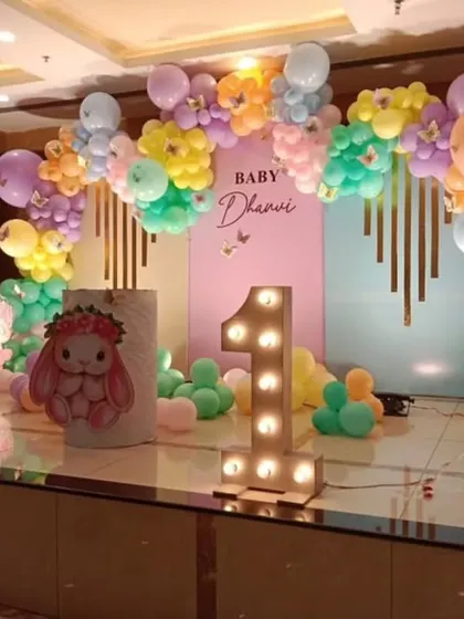 A cute bunny-themed first birthday party. The stage decor includes a pastel balloon arch, a pink backdrop, and a light-up marquee number '1'.