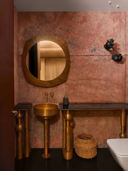 A bathroom at the Baid Residence featuring walls of red travertine, a freestanding brass washbasin by Vikram Goyal, and Bocci lights. The combination of the richly colored, porous stone and the gleaming, hand-beaten metal creates a luxurious and tactile experience.