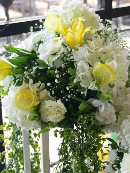 A beautiful floral arrangement featuring white hydrangeas, yellow lilies, and lemon slices. The cascading greenery adds a touch of drama and elegance to the citrus theme.