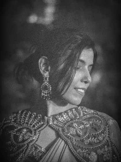 A soft, smiling portrait in black and white, focusing on the intricate details of the embroidered cape and earrings with a retro aesthetic.