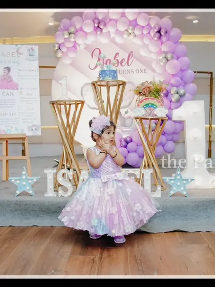The birthday girl, Isabel, looks precious in front of her baby elephant-themed first birthday decor. The setup uses a soft lavender and white color palette with cute elephant illustrations and "BABY" blocks.
