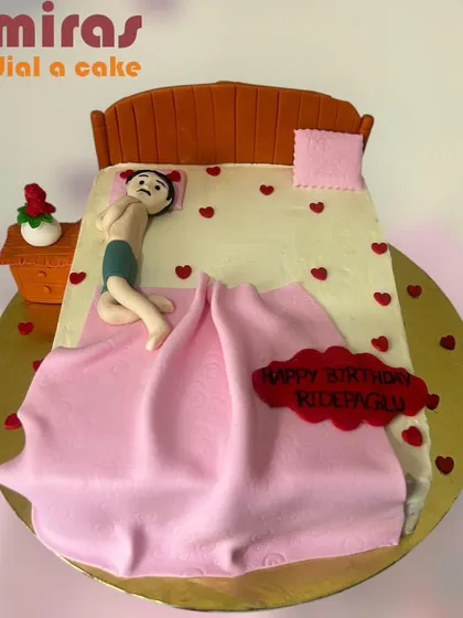 A funny and personalized cake showing a person relaxing in bed. This sculpted cake is a playful nod to someone who loves their comfort, complete with a tiny rose on the nightstand.