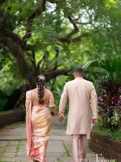 A candid shot from behind shows the beautiful fit and fall of the groom's pastel pink sherwani, coordinating perfectly with his bride's saree as they walk through a garden.