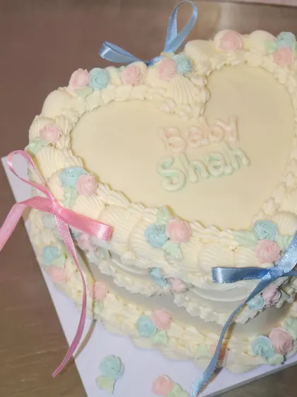 A sweet heart-shaped cake for a baby shower, decorated in a classic vintage style. We used pastel pink and blue buttercream rosettes and delicate ribbons to create this adorable centerpiece.