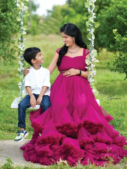 A serene moment between a mother-to-be and her son on a floral-decorated swing. The mother, in a stunning magenta gown, shares a loving gaze with her child, showcasing a peaceful and picturesque side of our outdoor maternity sessions.
