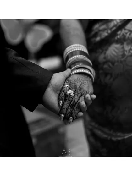 A close-up detail shot in black and white, focusing on the couple holding hands. The bride's intricate henna and bangles stand out, symbolizing their union.
