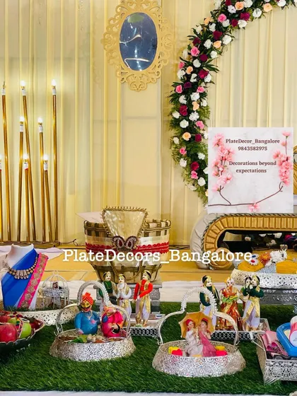 An elegant wedding gift display featuring Marapachi Bommai, saree platters, and other decorated items, all arranged in front of the stage.