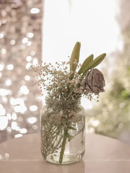 Sometimes, simple is most beautiful. This minimalist arrangement of a single silver-dusted rose and baby's breath in a mason jar adds a delicate and charming touch to a tablescape, with a soft, sparkling background.