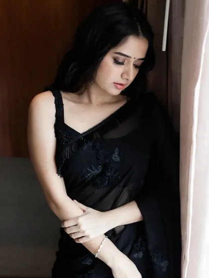 A moody portrait showcasing the delicate details of the black georgette saree, including the fine embroidery and the handmade tassels on the strap of the blouse.