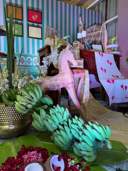 A whimsical corner featuring a decorative wooden horse, traditional banana offerings, and a backdrop of blue and white stripes.