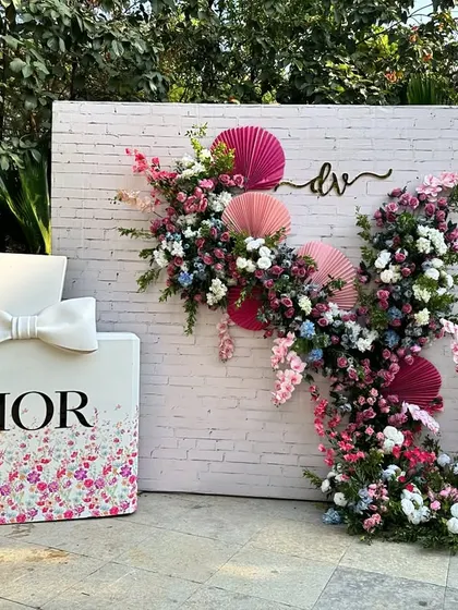 A Dior-themed photo booth with a white brick wall backdrop, adorned with a cascade of colorful flowers and paper fans, creating a perfect spot for stylish photos.