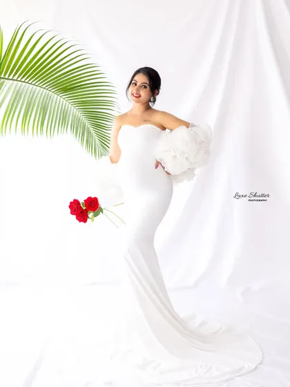 A full-length view showcasing the beautiful silhouette of the mother-to-be in a form-fitting white gown. The ruffled sleeves and pop of red from the roses add a high-fashion feel.