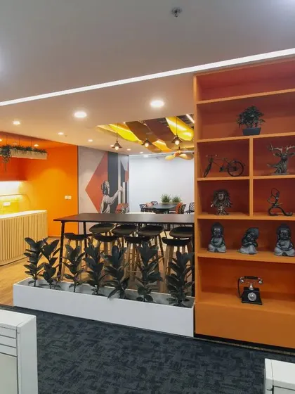 This view shows the transition from the workspace to the canteen, with a decorative shelving unit acting as a subtle divider. The high-top table and stools offer a casual spot for employees to eat or work.