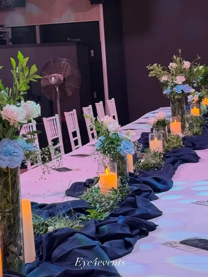 Another angle of the blue and white themed dining setup, showing the arrangement of candles and floral vases. The white Chiavari chairs add a touch of elegance to the overall party decoration.