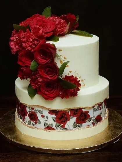 This two-tier wedding cake features a cascade of fresh red roses and carnations. The bottom tier is wrapped in an edible print with a matching rose pattern, adding a unique and personal touch.